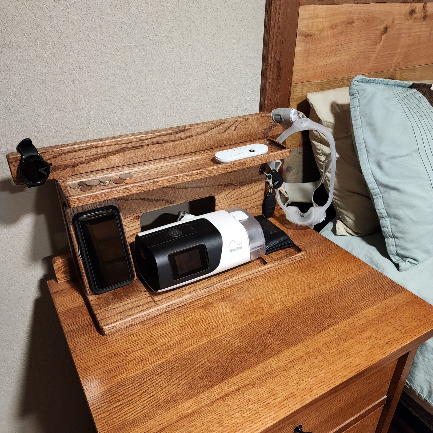 A CPAP Smart Stand that is displayed next to a bed holding the CPAP machine, wirelessly charging the phone and watch and has a few items in the catch-all trays.  The mask is also hanging from the stand to keep it off the nightstand and floor.  Items on the stand are very organized.  