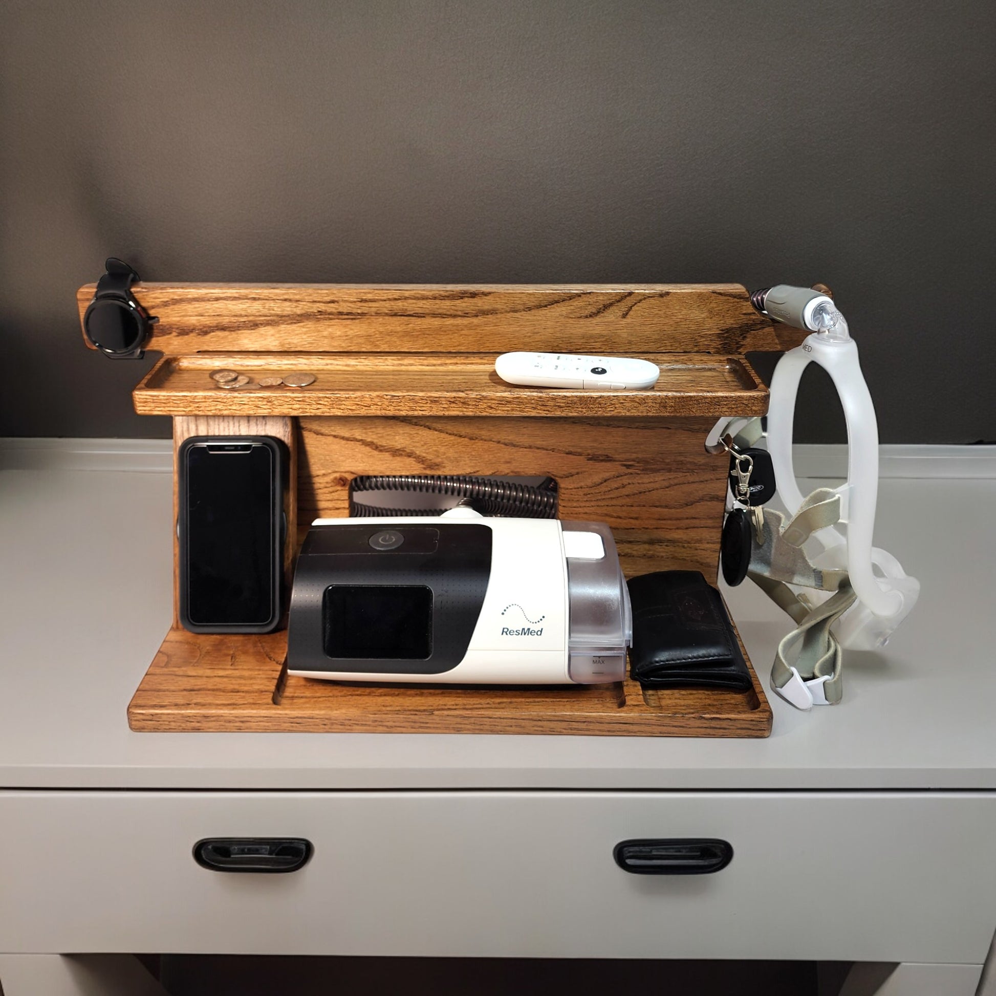 A CPAP Smart Stand that is charging a smart watch and charging a smart phone.  The stand is holding the CPAP machine, change, TV remote control, car keys and a wallet.  The mask is hanging from the stand to keep it off the nightstand and floor.  The stand has three catch-all trays and is made from solid oak wood.