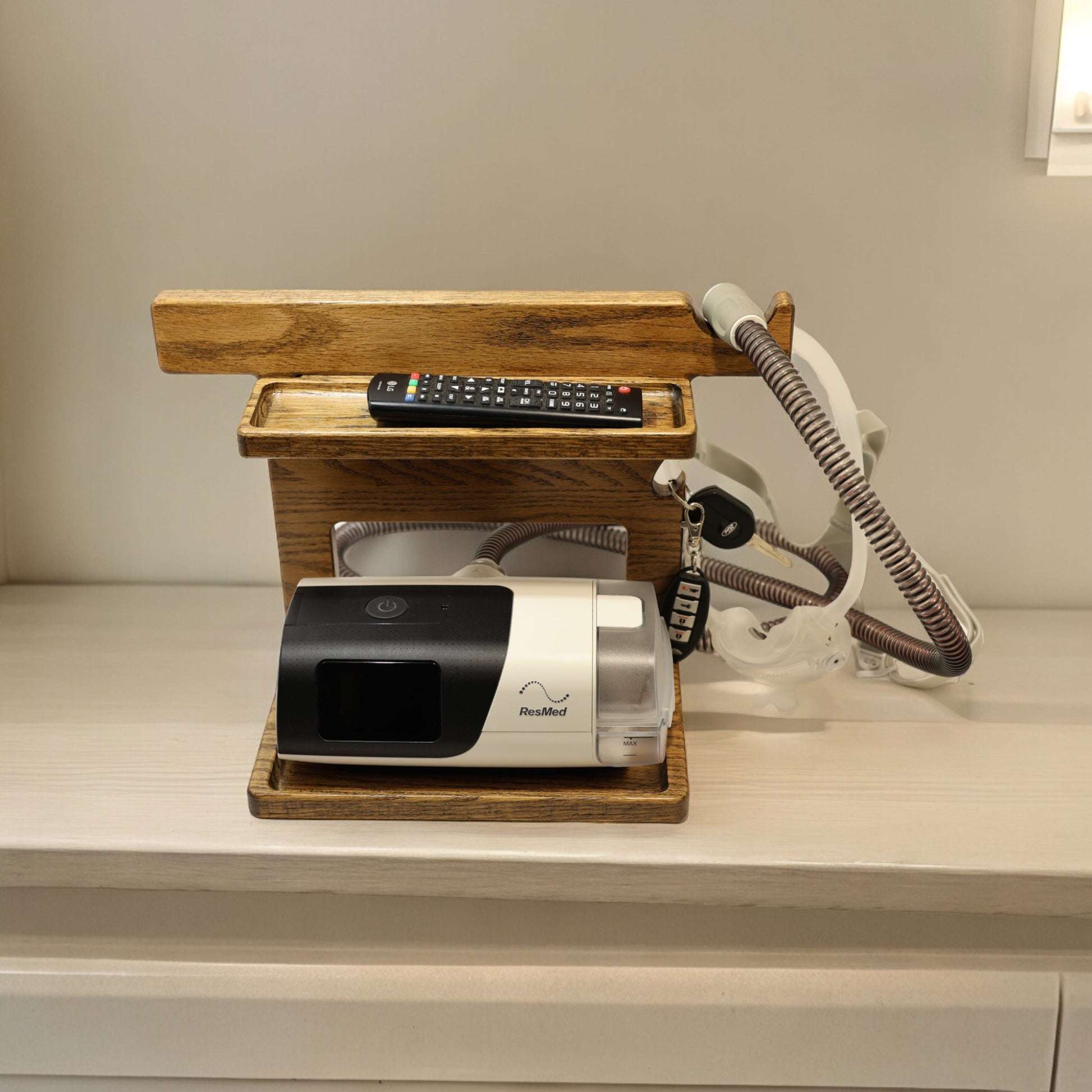 A CPAP Stand that has the CPAP inserted and mask hanging from the stand. The stand is made by Custom Cave works and is made from solid oak wood.  The stand is also holding car keys and a TV remote. 