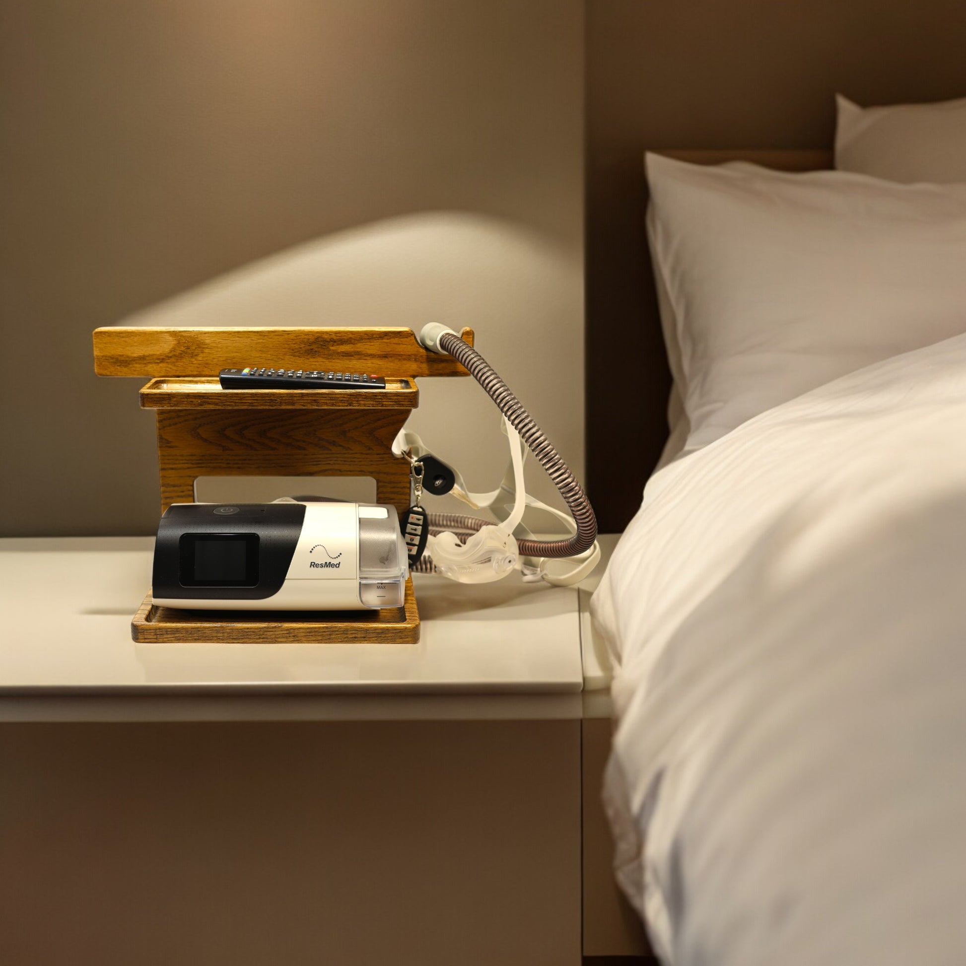 A CPAP Stand next to the comfortable bed,  The stand is holding the CPAP machine, TV remote control, and has room for other items.  The mask is hanging from the stand to keep it off the nightstand and floor.  The stand has two catch-all trays and is made from solid oak wood.